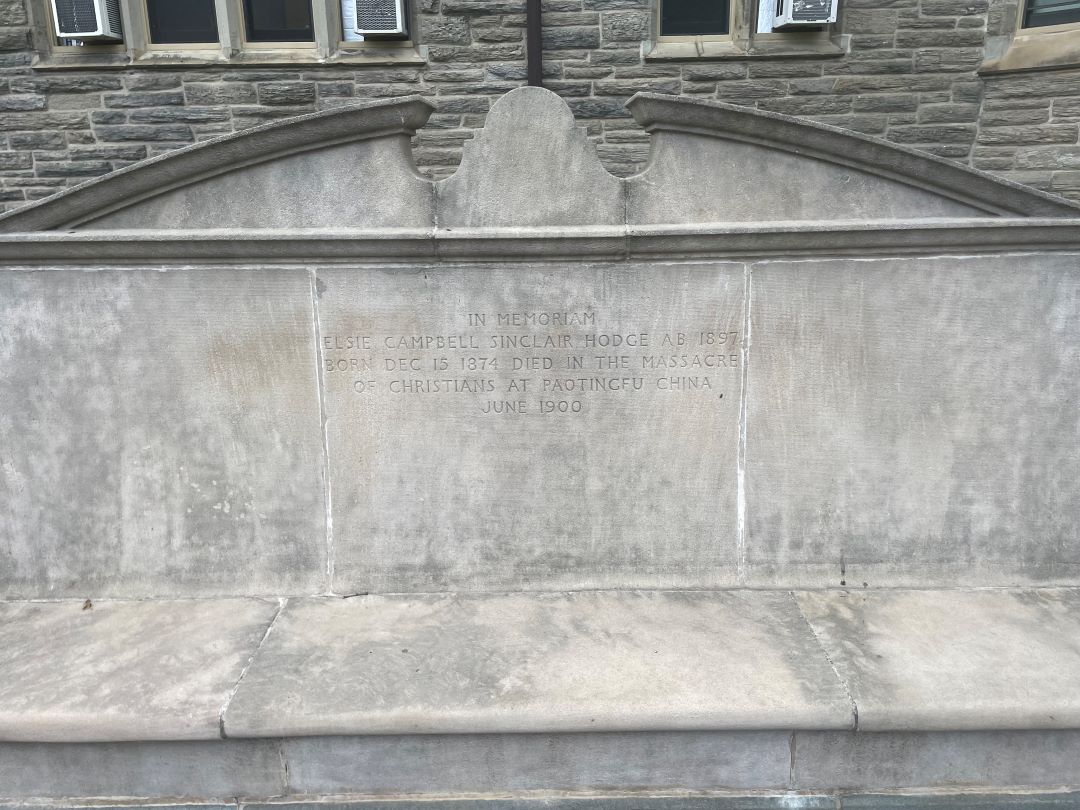 A gray, concrete bench between Denbigh and Guild Halls. The inscription on the bench is In Memoriam Elsie Campbell Sinclair Hodge A.B. 1897 Born December 1874 Died in the massacre of Christians at Paotingfu China June 1900