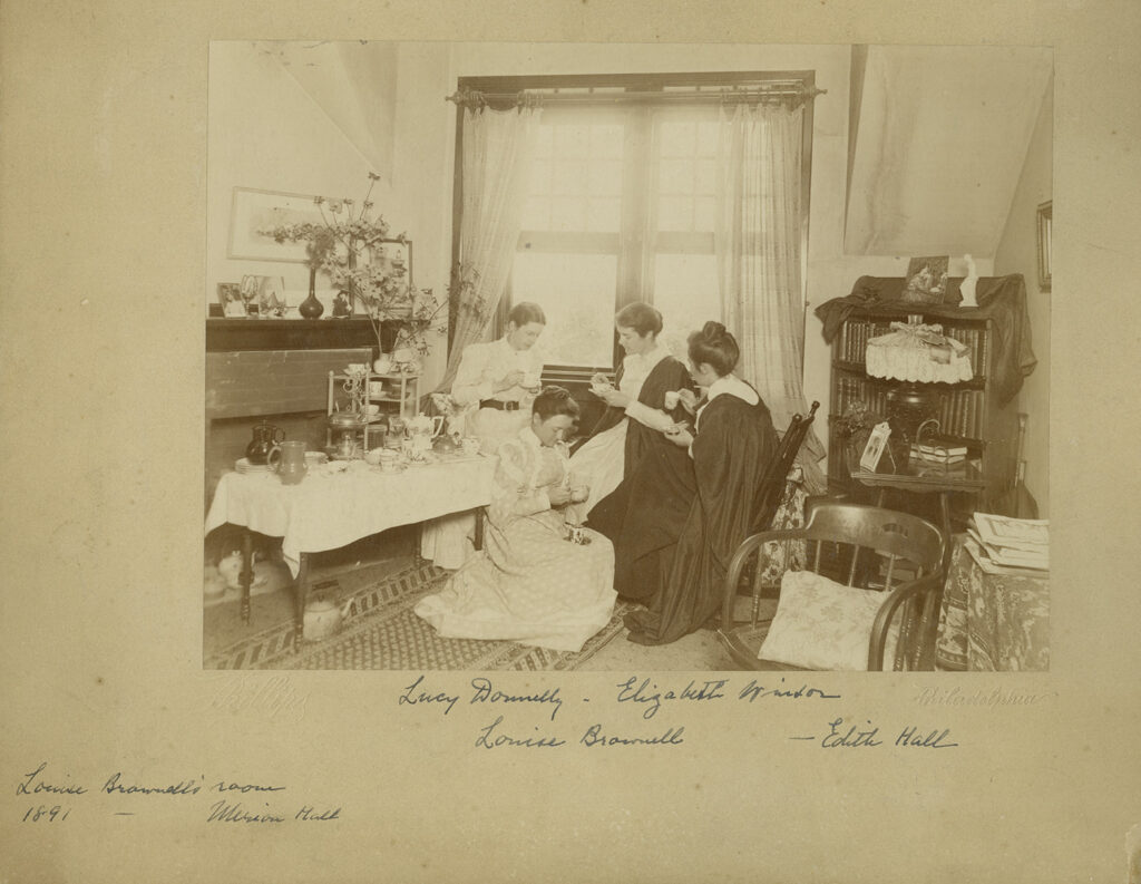 Women sit in an adorned dorm room, having tea. Some of the women wear black robes. 