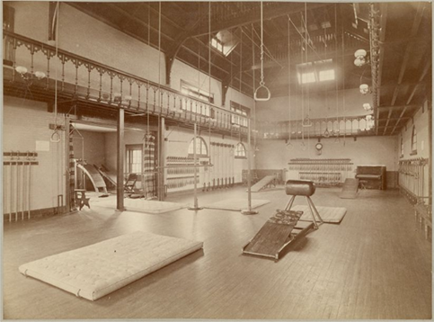 Interior view of the old gymnasium. The image depicts a corner of a gym room filled with athletic equipment. Several mats and ramps lay on the floor. Several weights and bats are hung on the walls. The photograph is matted and the back of the mat contains another photograph of a graveyard. The photograph is from the Phillips Photograph Album (1899).
