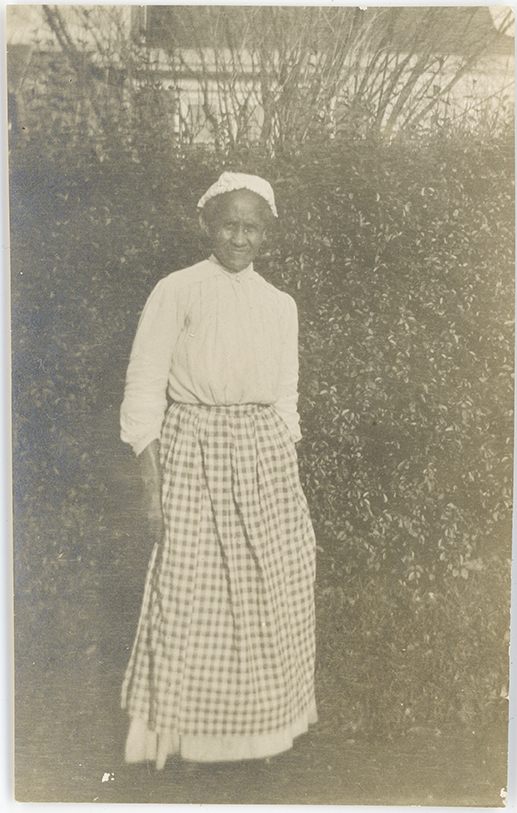 An unnamed maid stands outside. She wears a plain dress with a checkered apron and a plain head covering. 