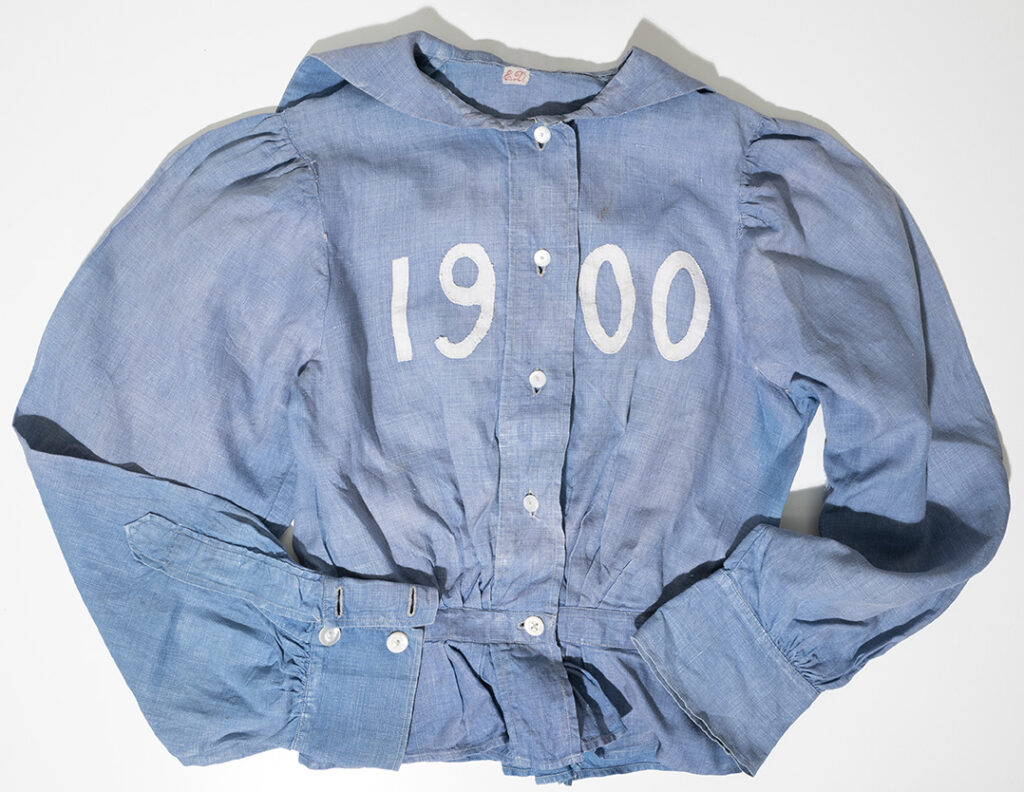 A student's gym blouse. The blouse is on blue cotton with the year 1900 across the front in white. 