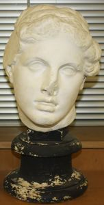 A cast of a classical bust. It is mounted on a black pedestal and the nose of the bust is damaged. 