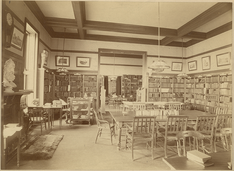A photograph of the interior of a room in Taylor Hall that served as a library room. Framed picture line the walls and shelves wrap around the room alongside the wall. Tables and chairs are positioned in the middle of the room for studying. The side of the fireplace can be seen. 