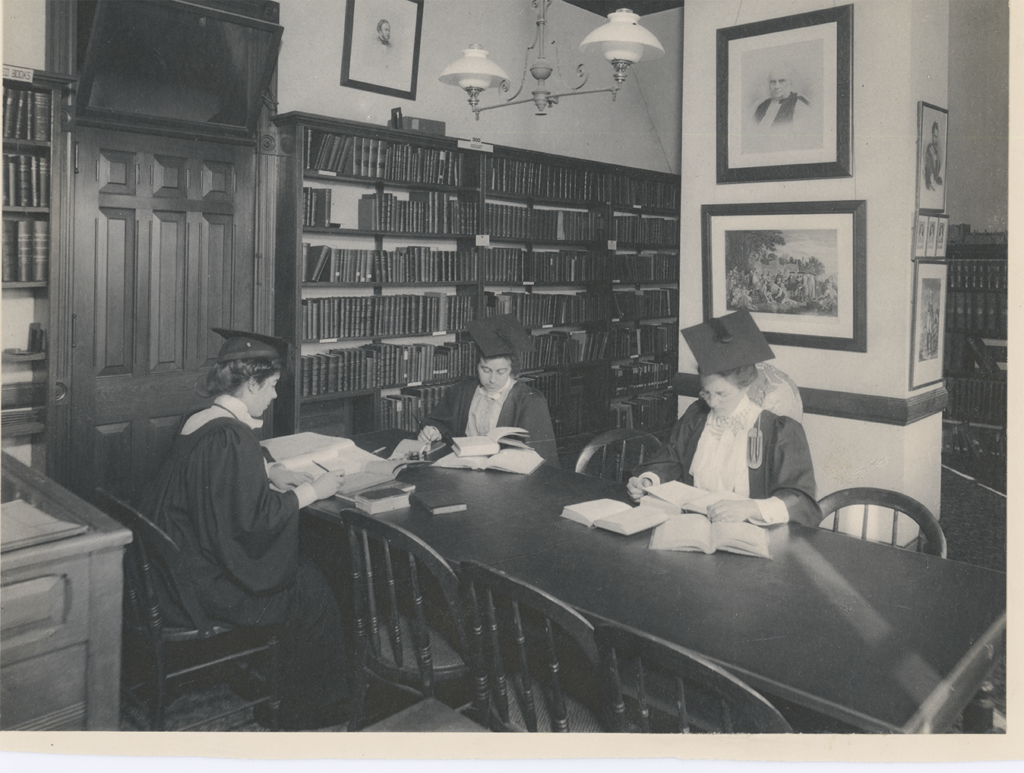 Photograph of Bryn Mawr students studying in Taylor Hall Library. Three students sit around a table and read while wearing black gowns and caps. A framed picture of William Penn signing a treaty with the region's Indigenous Peoples can be seen on the wall behind the students.